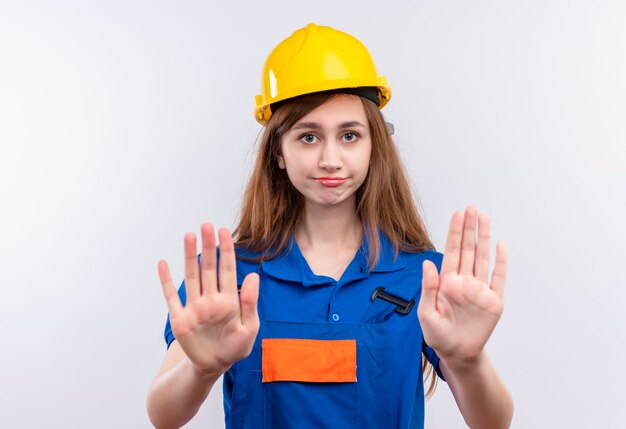 Trabajador constructor joven en uniforme de construcción y casco de seguridad mirando disgustado de pie con las manos abiertas haciendo señal de stop sobre pared blanca