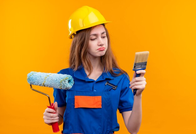 Trabajador constructor joven en uniforme de construcción y casco de seguridad sosteniendo cepillo y rodillo de pintura mirando cepillo con expresión escéptica de pie sobre pared naranja