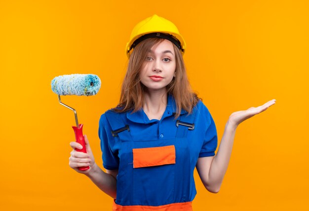 Trabajador constructor de mujer joven en uniforme de construcción y casco de seguridad con rodillo de pintura mirando confundido encogiéndose de hombros de pie sobre la pared naranja