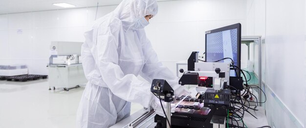 Un trabajador de fábrica con un traje de laboratorio blanco, guantes de látex y máscara facial que trabaja con algunos equipos modernos durante la fabricación de un televisor