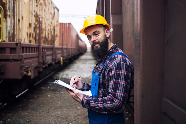 Trabajador del ferrocarril con portapapeles de pie junto a los contenedores de envío y mirando hacia el frente