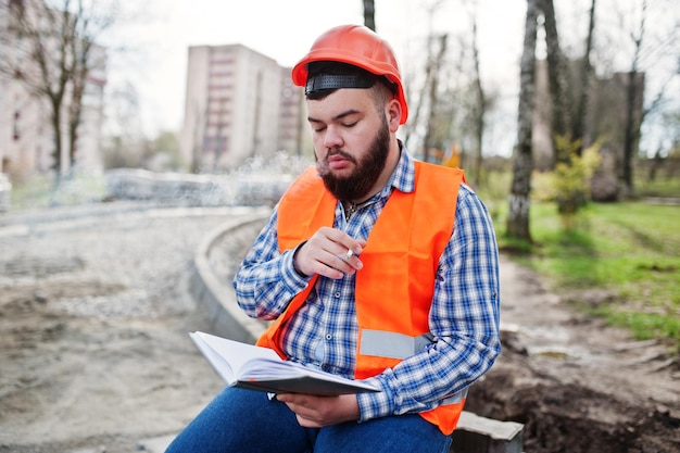 Trabajador fumador de barba traje de hombre trabajador de la construcción en casco naranja de seguridad sentado en el descanso del pavimento en el trabajo y leyendo las entradas del cuaderno de trabajo