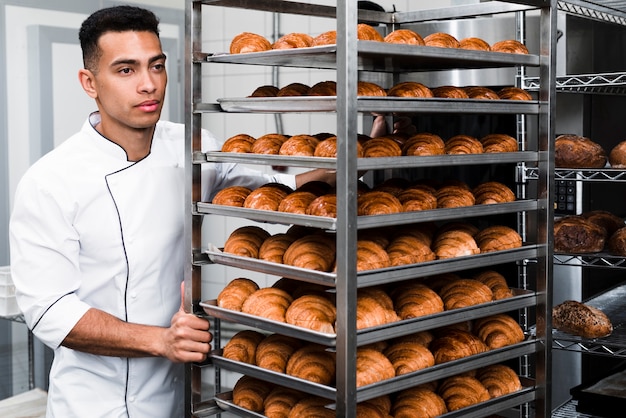 Trabajador guapo en uniforme llevando estantes con croissant en la panadería