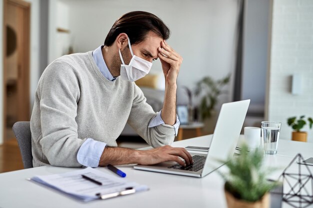 Trabajador independiente preocupado con mascarilla protectora mientras lee correos electrónicos problemáticos en una computadora y trabaja en casa durante la epidemia de virus
