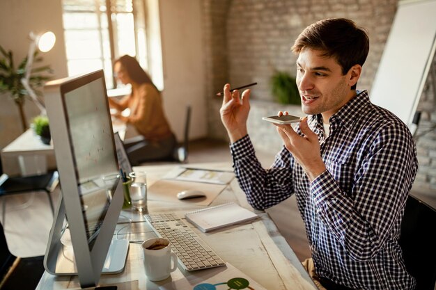 Trabajador independiente sonriente comunicándose por el altavoz del teléfono móvil mientras lee informes en una computadora en la oficina