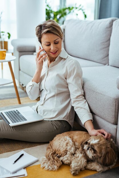 Trabajador independiente sonriente con un perro que se comunica por teléfono celular mientras trabaja en una computadora portátil en casa