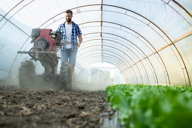 Trabajador joven agricultor que opera el motor cultivador para preparar el suelo para nuevas plántulas en una granja de alimentos orgánicos