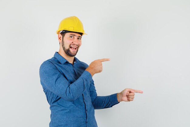 Trabajador joven apuntando hacia el lado en camisa, casco y mirando alegre.