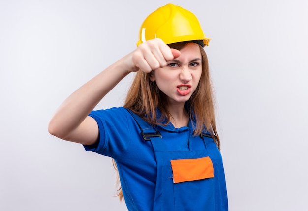 Trabajador joven constructor en uniforme de construcción y casco de seguridad apretando el puño en la parte delantera