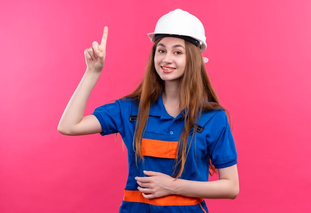 Trabajador joven constructor en uniforme de construcción y casco de seguridad apuntando con el dedo hacia arriba con gran idea sonriendo de pie sobre la pared rosa