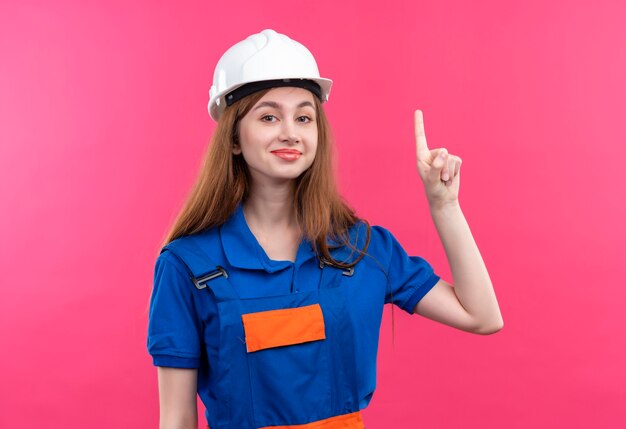 Trabajador joven constructor en uniforme de construcción y casco de seguridad apuntando con el dedo hacia arriba con gran idea sonriendo de pie sobre la pared rosa