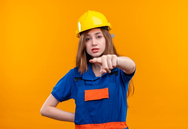 Trabajador joven constructor en uniforme de construcción y casco de seguridad apuntando con el dedo índice al frente disgustado de pie sobre la pared naranja