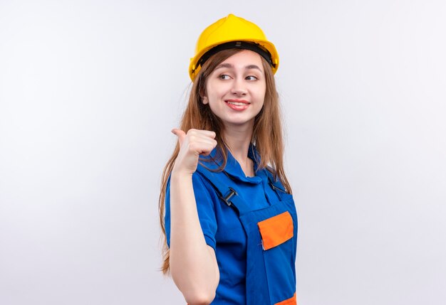 Trabajador joven constructor en uniforme de construcción y casco de seguridad apuntando con el pulgar hacia el lado sonriendo confiado de pie sobre la pared blanca