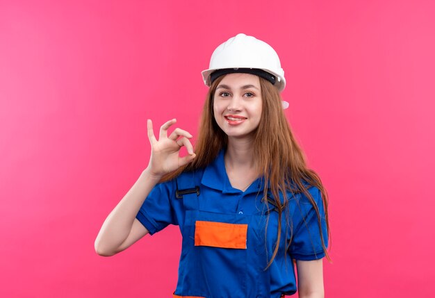 Trabajador joven constructor en uniforme de construcción y casco de seguridad feliz y positivo sonriendo mostrando signo ok de pie sobre pared rosa