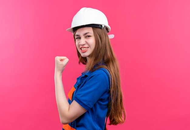 Trabajador joven constructor en uniforme de construcción y casco de seguridad levantando el puño mirando confiado, posando como un ganador de pie sobre una pared rosa