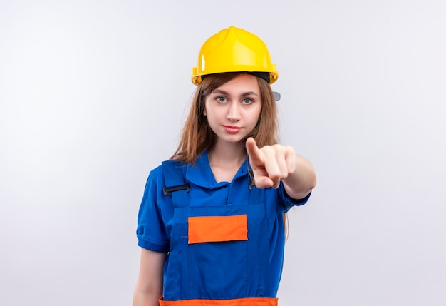 Trabajador joven constructor en uniforme de construcción y casco de seguridad mirando con cara seria apuntando con el dedo índice