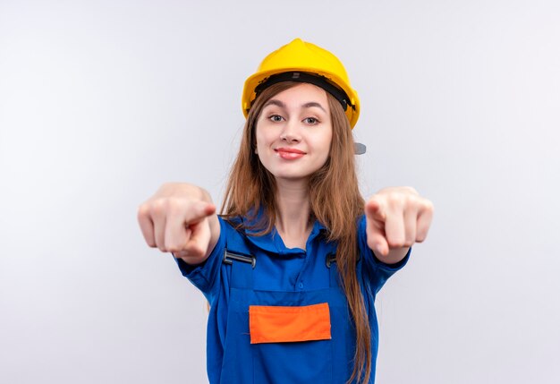 Trabajador joven constructor en uniforme de construcción y casco de seguridad mirando confiado sonriendo apuntando con los dedos índices en la parte delantera