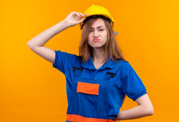 Trabajador joven constructor en uniforme de construcción y casco de seguridad mirando disgustado, frunciendo el ceño tocando su casco de pie