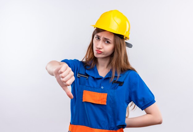 Trabajador joven constructor en uniforme de construcción y casco de seguridad mirando disgustado mostrando los pulgares hacia abajo de pie sobre la pared blanca