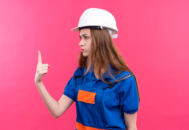 Trabajador joven constructor en uniforme de construcción y casco de seguridad mirando a un lado mostrando el dedo índice con el ceño fruncido de pie sobre la pared rosa