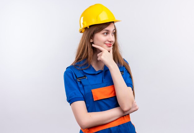 Trabajador joven constructor en uniforme de construcción y casco de seguridad mirando a un lado de pie con la mano en la barbilla con expresión pensativa pensando sobre la pared blanca