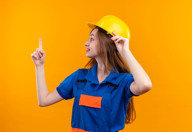 Trabajador joven constructor en uniforme de construcción y casco de seguridad mirando a un lado con una sonrisa en la cara mostrando el dedo índice de pie