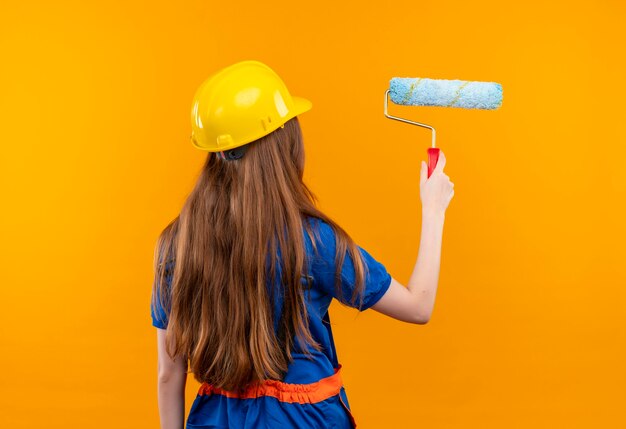 Trabajador joven constructor en uniforme de construcción y casco de seguridad de pie con la espalda yendo a pintar con rodillo de pintura sobre pared naranja