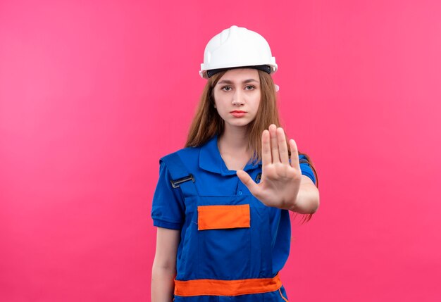 Trabajador joven constructor en uniforme de construcción y casco de seguridad de pie con la mano abierta haciendo señal de stop