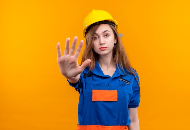 Trabajador joven constructor en uniforme de construcción y casco de seguridad de pie con la mano abierta haciendo señal de stop