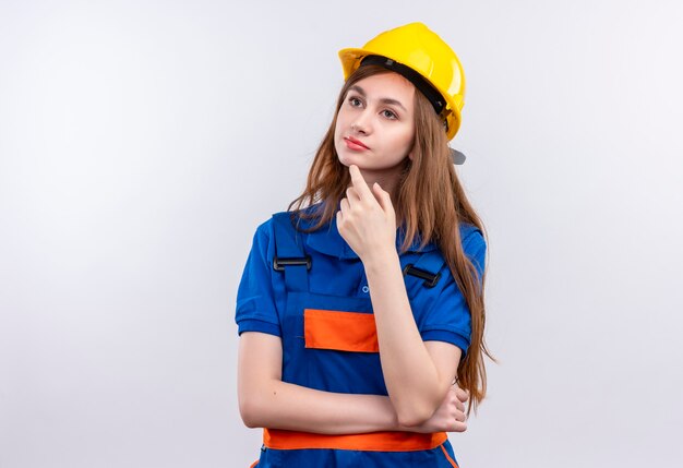 Foto gratuita trabajador joven constructor en uniforme de construcción y casco de seguridad de pie con la mano en la barbilla con expresión pensativa pensando sobre la pared blanca