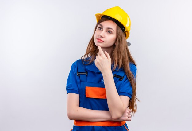 Trabajador joven constructor en uniforme de construcción y casco de seguridad de pie con la mano en la barbilla con expresión pensativa pensando sobre la pared blanca