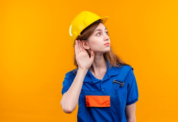 Trabajador joven constructor en uniforme de construcción y casco de seguridad de pie con la mano cerca de la oreja tratando de escuchar de pie sobre la pared naranja