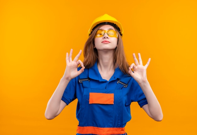 Trabajador joven constructor en uniforme de construcción y casco de seguridad de pie con los ojos cerrados relajándose haciendo gesto de meditación con los dedos sobre la pared naranja