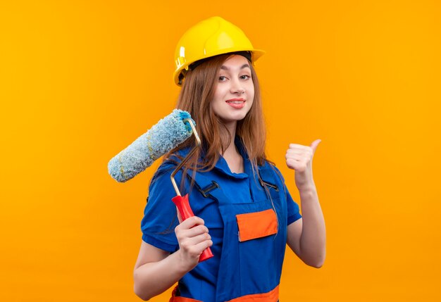 Trabajador joven constructor en uniforme de construcción y casco de seguridad con rodillo de pintura mostrando los pulgares para arriba sonriendo confiado de pie sobre la pared naranja