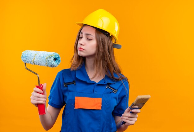 Trabajador joven constructor en uniforme de construcción y casco de seguridad con rodillo de pintura y pincel mirando el rodillo con expresión escéptica de pie sobre la pared naranja