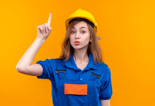 Trabajador joven constructor en uniforme de construcción y casco de seguridad, señalando con el dedo índice hacia arriba advertencia con cara seria de pie sobre la pared naranja