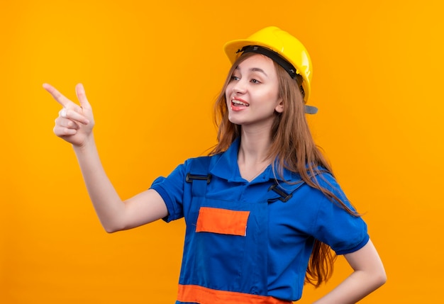 Trabajador joven constructor en uniforme de construcción y casco de seguridad sonriendo alegremente apuntando con el dedo índice hacia el lado parado sobre la pared naranja