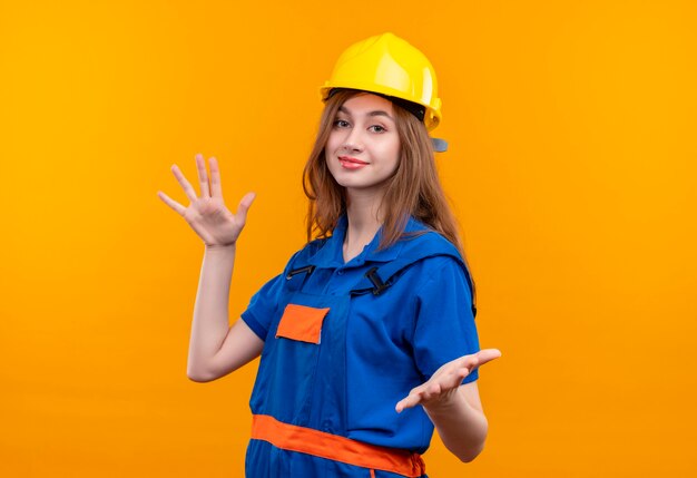 Trabajador joven constructor en uniforme de construcción y casco de seguridad sonriendo amable haciendo gesto de bienvenida con las manos abiertas de pie sobre la pared naranja