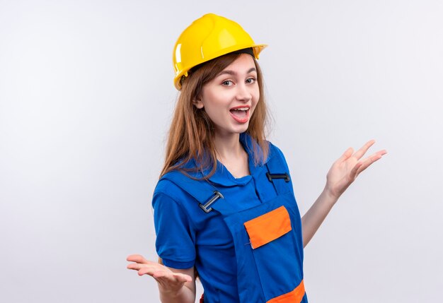 Trabajador joven constructor en uniforme de construcción y casco de seguridad sonriendo ampliamente extendiendo los brazos a los lados de pie sobre la pared blanca