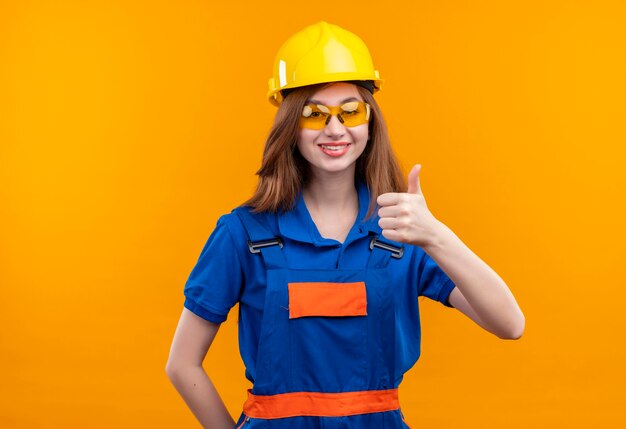 Trabajador joven constructor en uniforme de construcción y casco de seguridad sonriendo ampliamente mostrando los pulgares para arriba de pie sobre la pared naranja