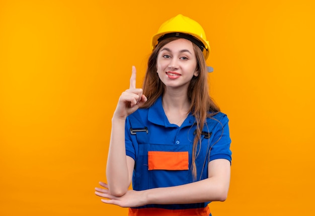 Trabajador joven constructor en uniforme de construcción y casco de seguridad sonriendo confiado apuntando con el dedo índice hacia arriba teniendo buena idea de pie sobre la pared naranja