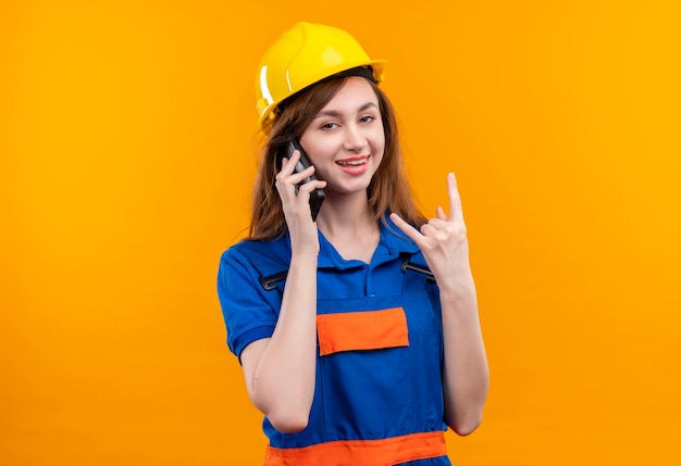 Trabajador joven constructor en uniforme de construcción y casco de seguridad sonriendo mientras habla por teléfono móvil, haciendo el símbolo de la roca con los dedos de pie sobre la pared naranja