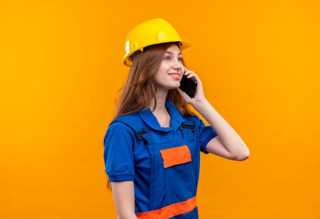 Trabajador joven constructor en uniforme de construcción y casco de seguridad sonriendo mientras habla por teléfono móvil de pie sobre la pared naranja