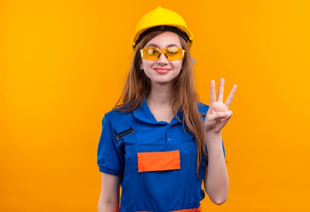 Trabajador joven constructor en uniforme de construcción y casco de seguridad sonriendo mostrando y apuntando hacia arriba con los dedos número tres de pie sobre la pared naranja