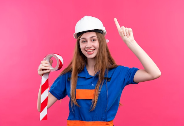 Trabajador joven constructor en uniforme de construcción y casco de seguridad sosteniendo cinta adhesiva apuntando con el dedo índice hacia arriba sonriendo con gran idea de pie sobre la pared rosa