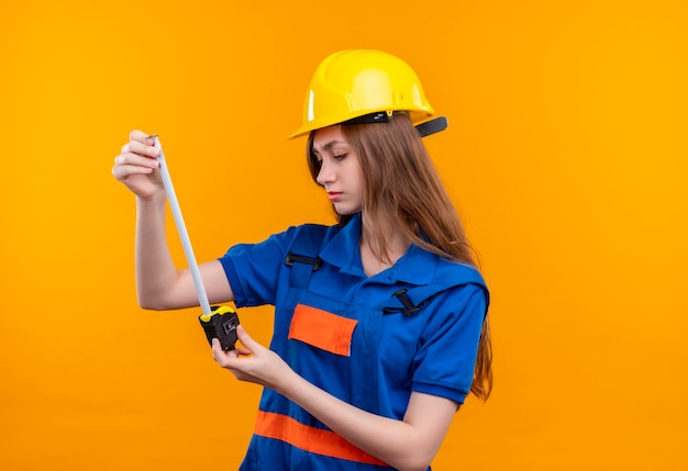 Trabajador joven constructor en uniforme de construcción y casco de seguridad sosteniendo cinta métrica mirándolo con cara seria de pie sobre pared naranja