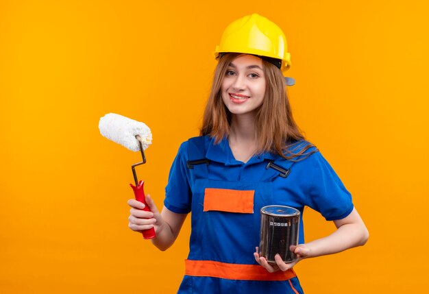 Trabajador joven constructor en uniforme de construcción y casco de seguridad sosteniendo lata de pintura y rodillo sonriendo