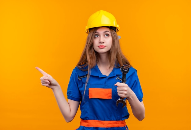 Trabajador joven constructor en uniforme de construcción y casco de seguridad sosteniendo una llave apuntando con el dedo índice hacia el lado