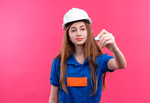 Trabajador joven constructor en uniforme de construcción y casco de seguridad tratando de escribir algo en el aire con lápiz parado sobre pared rosa
