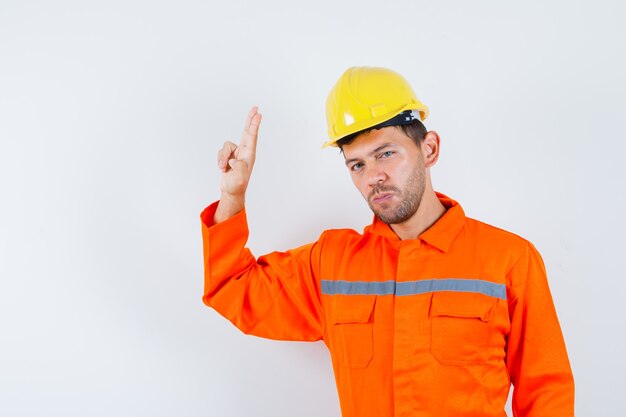 Trabajador joven gesticulando con la mano y los dedos en uniforme, casco y mirando confiado.